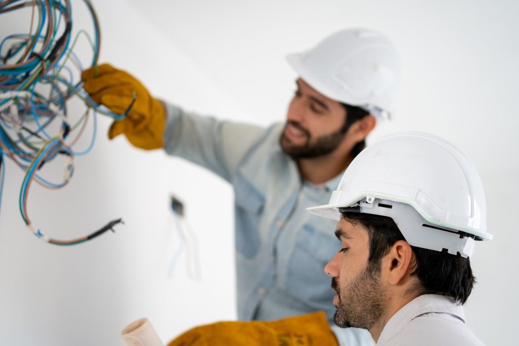 Two electricians in branded uniforms working inside a Dubai apartment, while a family relaxes with lights on in the background.