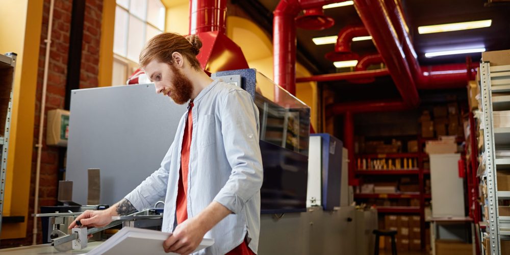 Bearded Man Working at Print Factory Side view portrait of bearded young man operating industrial printing machine at workshop, copy space
