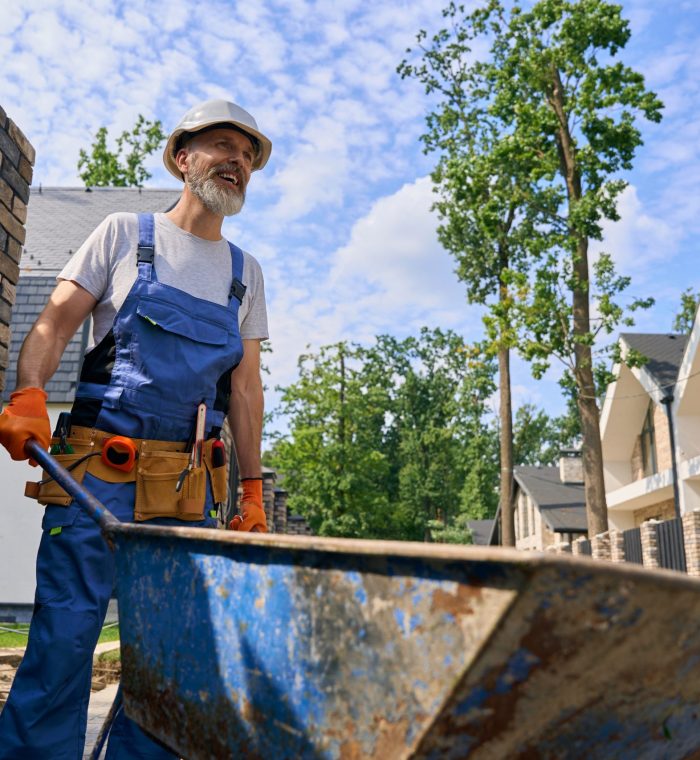 Joyous builder transferring building materials on site in morning Cheerful construction worker in hard hat and rubber gloves pushing wheelbarrow forward among unfinished houses