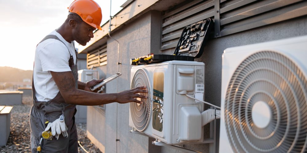 Male holding tablet and installing cooling system outdoors Qualified african american engineer looking at hanging air conditioner on rooftop of residential building. Competent craftsman holding modern tablet and installing device step by step outdoors.